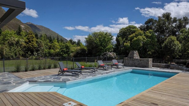 Piscina retangular com deck de madeira, cercada por um muro de pedra, e três cadeiras de descanso com almofadas vermelhas.