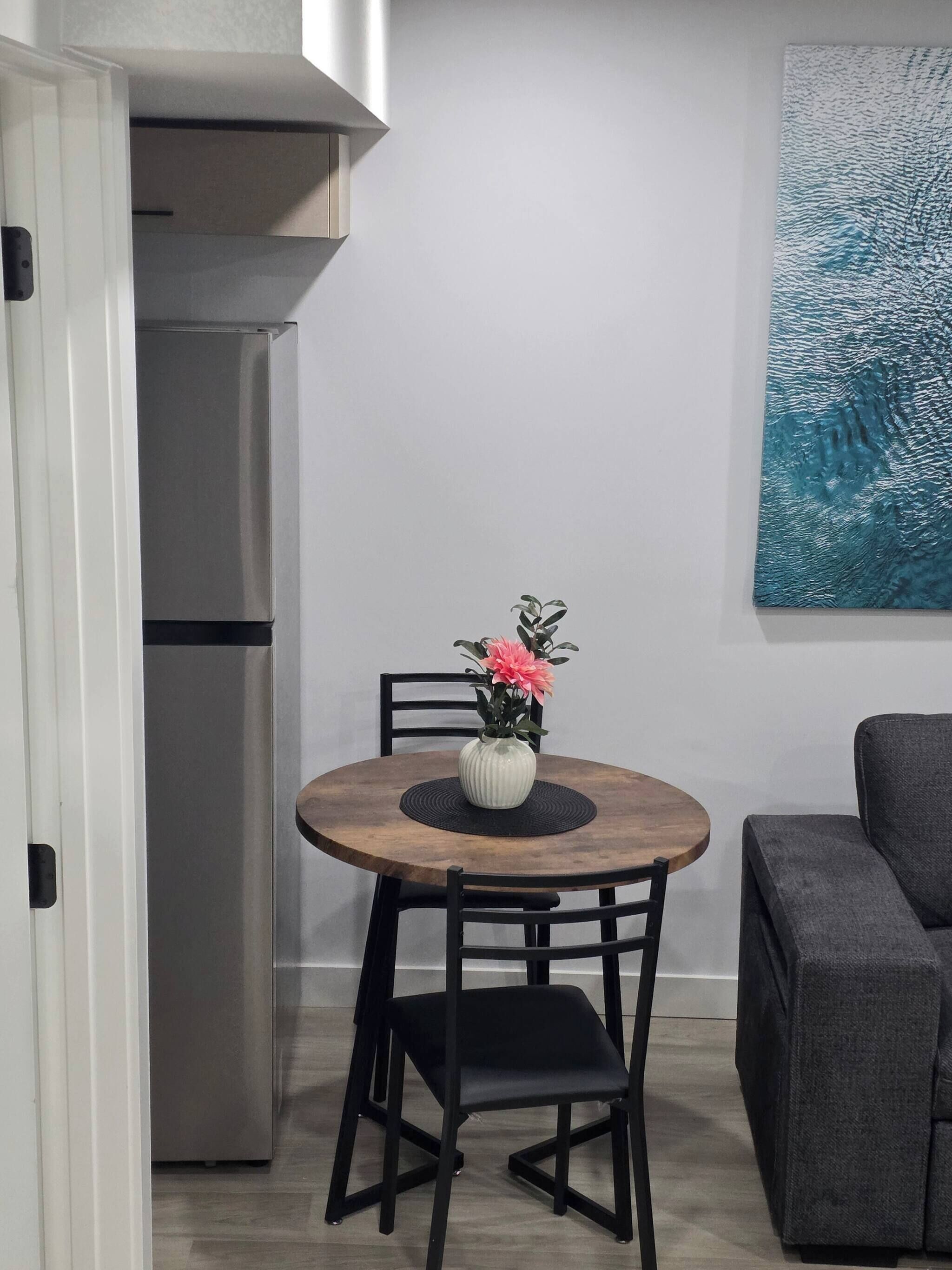 Une cuisine compacte équipée d’un réfrigérateur intégré, d’une table ronde en bois avec deux chaises et d’un tableau accroché au mur.