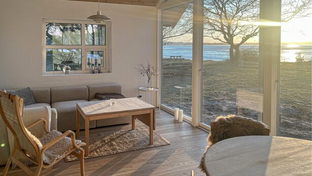 A cozy living room with a sofa, chairs, a table, and a large window with a view of a beach and sunset. - Amazing home in Skanderborg with sauna (Skanderborg)