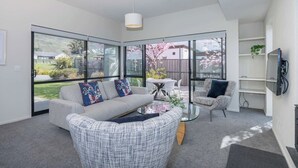 A modern living room with a sofa, armchair, glass coffee table, and a TV mounted on the wall. - Mountain views in Meadowstone (Wānaka)