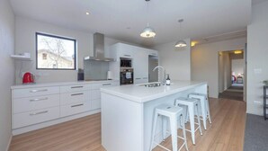 A modern kitchen with white cabinetry, a central island with bar stools, a window with a view, and a visible hallway leading to another room. - Mountain views in Meadowstone (Wānaka)