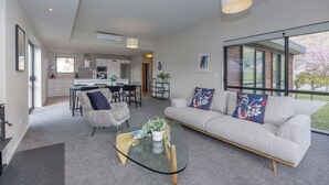 A modern living room with a glass coffee table, a sofa with decorative pillows, a dining area with chairs, and a kitchen in the background. - Mountain views in Meadowstone (Wānaka)