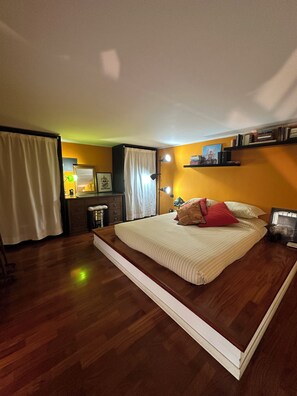 Ein Schlafzimmer mit einem großen Bett, Holzboden, einem Nachttisch, einer Lampe und einem Bücherregal.
