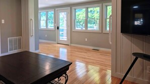 An open-plan living area with a dark wooden table, a television mounted on the wall, and large windows allowing natural light.