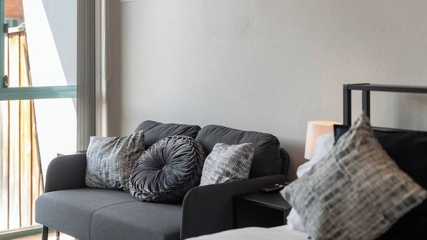 Ein modernes Wohnzimmer mit einer Couch und einem Bett, beide mit grauer Polsterung, und einem großen Fenster, das natürliches Licht hereinlässt.