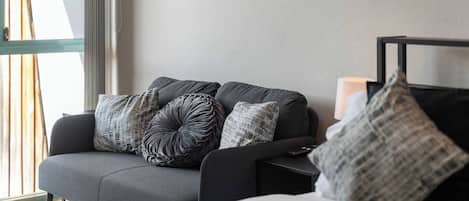 Un salon moderne comprenant un canapé et un lit, tous deux recouverts d’un tissu gris, ainsi qu’une grande fenêtre laissant entrer la lumière naturelle.