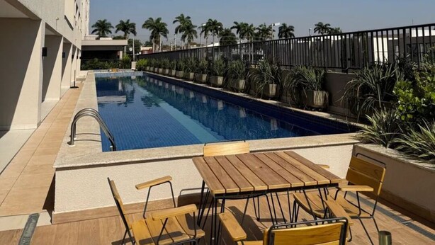 A swimming pool with a wooden deck and chairs, surrounded by a fence and palm trees. - 2-bedroom apartment, close to the 44th Street area, bus station, airport, and Flamboyant Shopping Mall. (Goiânia)