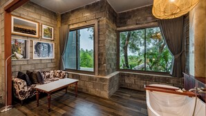 A room with a stone wall, a sofa, a table, a bathtub, and large windows with a view of greenery. - PhlaidamhillSichon (Sichon)