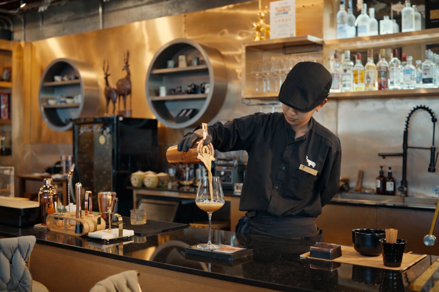 Un barista prépare une boisson au comptoir, derrière lequel on aperçoit des étagères remplies de bouteilles et, au fond, une sculpture décorative représentant un cerf.
