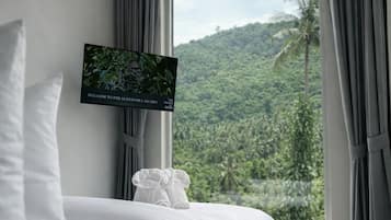 A modern bedroom with a large bed, a flat-screen TV mounted on the wall, and a view of a lush green landscape through a large window.
