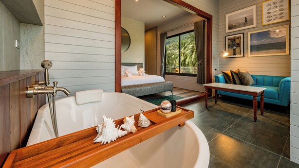 A modern bathroom with a bathtub, a wooden vanity, and a shower. There is a bed with a view of palm trees outside. - PhlaidamhillSichon (Sichon)
