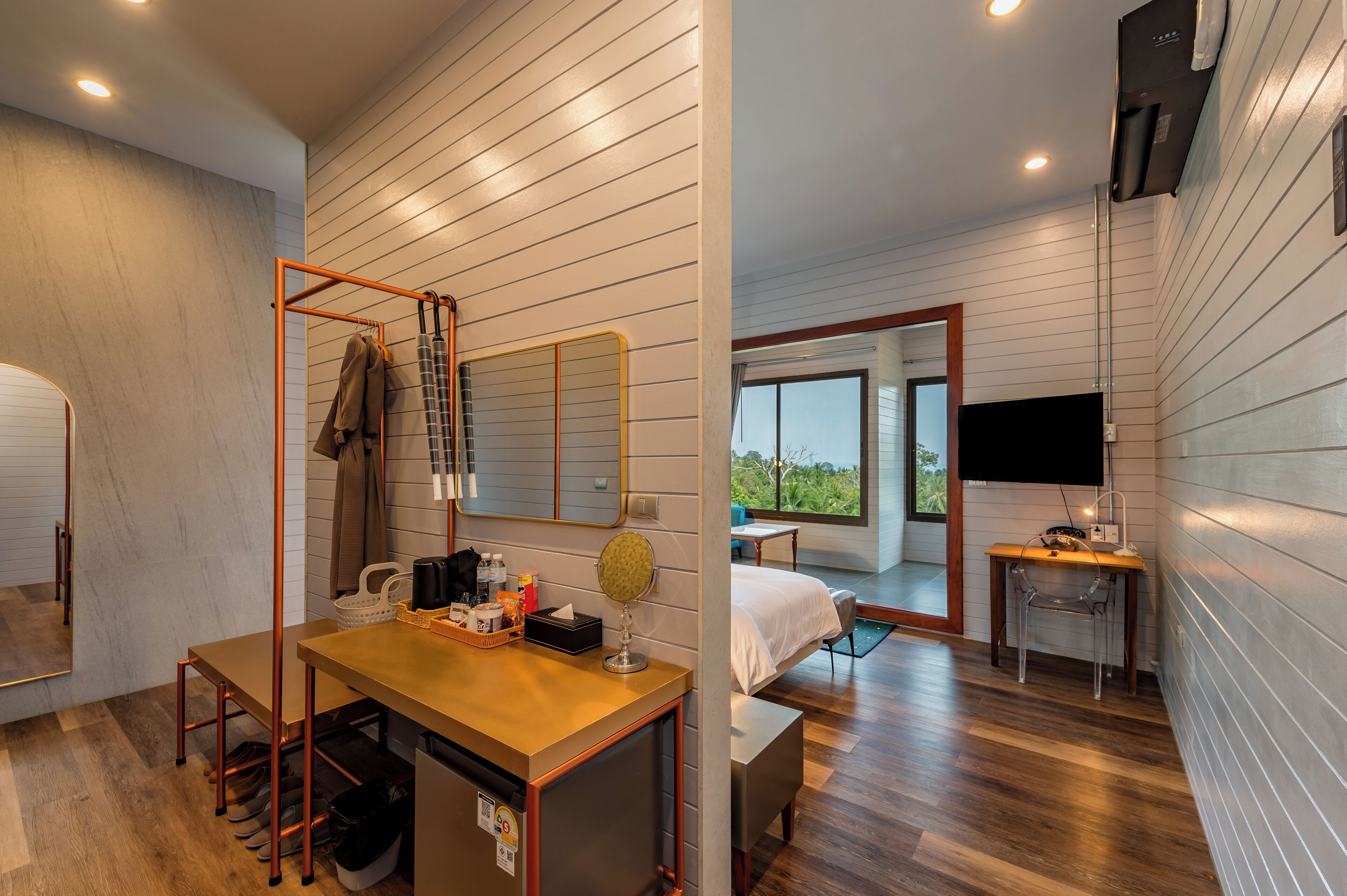 Habitación de hotel moderna con suelo de madera, un escritorio con silla, una cama, una mesita, un espejo y un televisor montado en la pared.