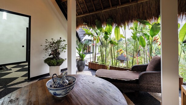 Une pièce avec une table en bois, un bol, une plante en pot, un canapé en osier et une vue sur une végétation luxuriante.