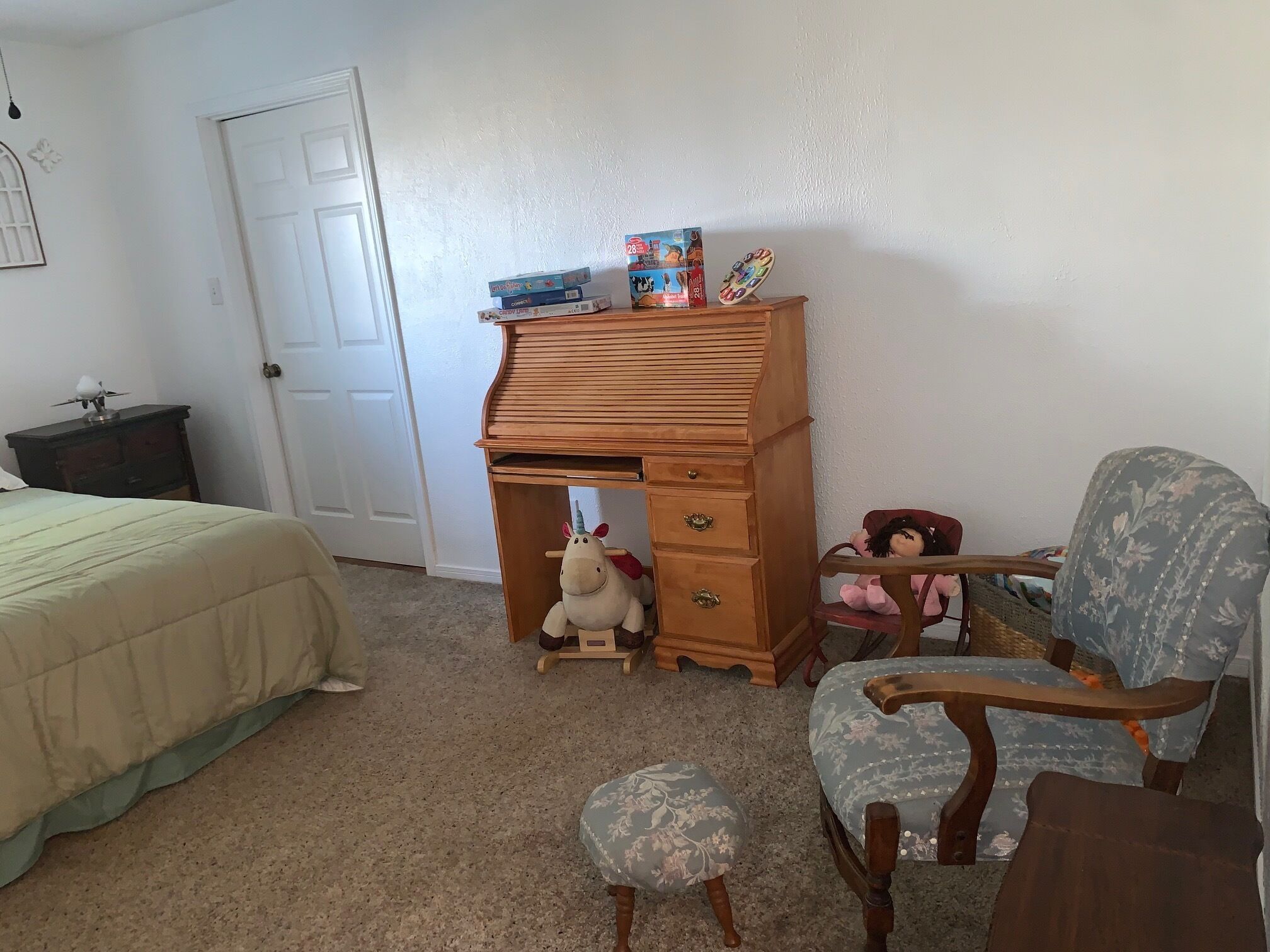 Un dormitorio con una cama, un escritorio de madera, una silla, un taburete pequeño y una puerta.