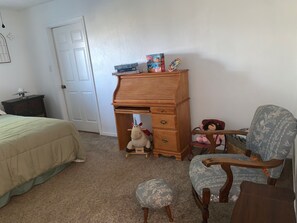 A bedroom with a bed, a wooden desk, a chair, a small stool, and a door.