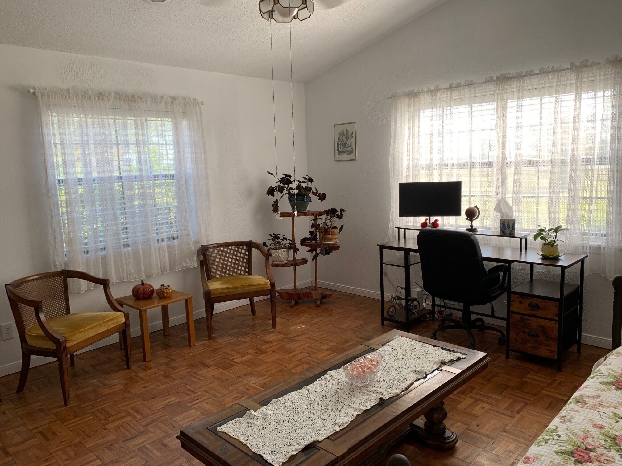 Una habitación con piso de madera, un escritorio con una computadora, dos sillas, una mesita con una planta y una cama con ropa de cama estampada.