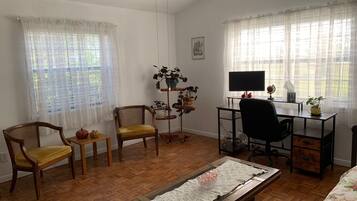 Una habitación con piso de madera, un escritorio con una computadora, dos sillas, una mesita con una planta y una cama con ropa de cama estampada.