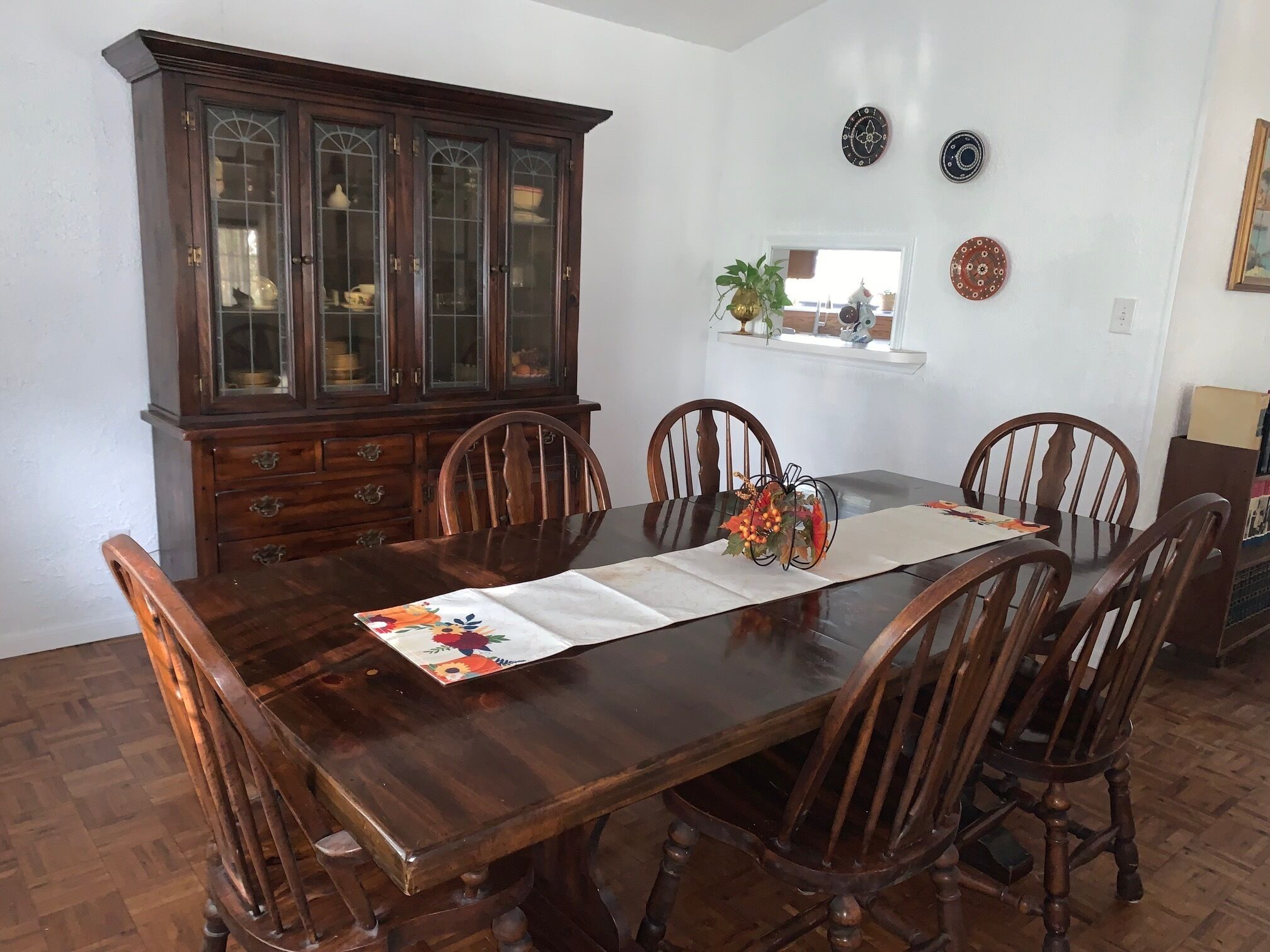 A dining room with a wooden table, six chairs, a wooden cabinet, and decorative plates on the wall.