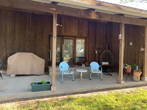 A covered patio with wooden walls, two blue chairs, a table, a barbecue cover, and potted plants.