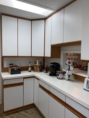 A kitchen with white cabinets, a countertop with snacks, a coffee maker, and a mug rack.