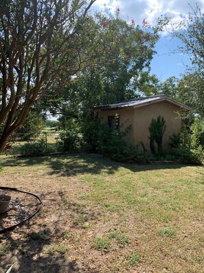 A small, single-story building with a sloped roof, surrounded by a grassy yard and dense foliage.