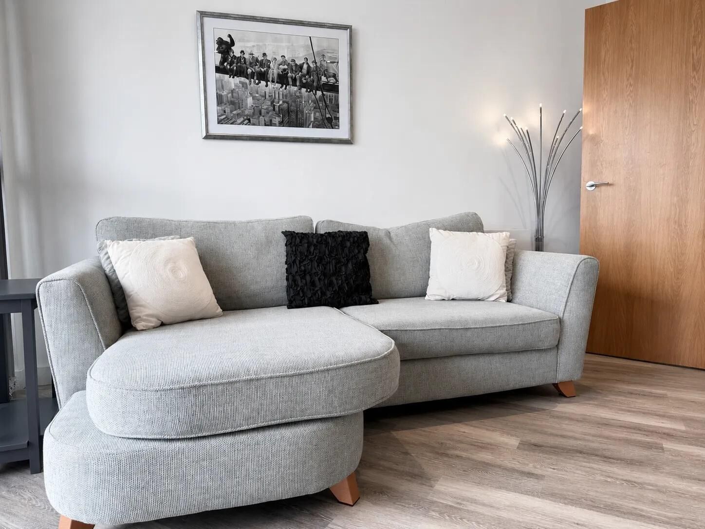 Ein modernes Wohnzimmer mit einem grauen Ecksofa, einem gerahmten Schwarz-Weiß-Foto an der Wand und einer Holztür.