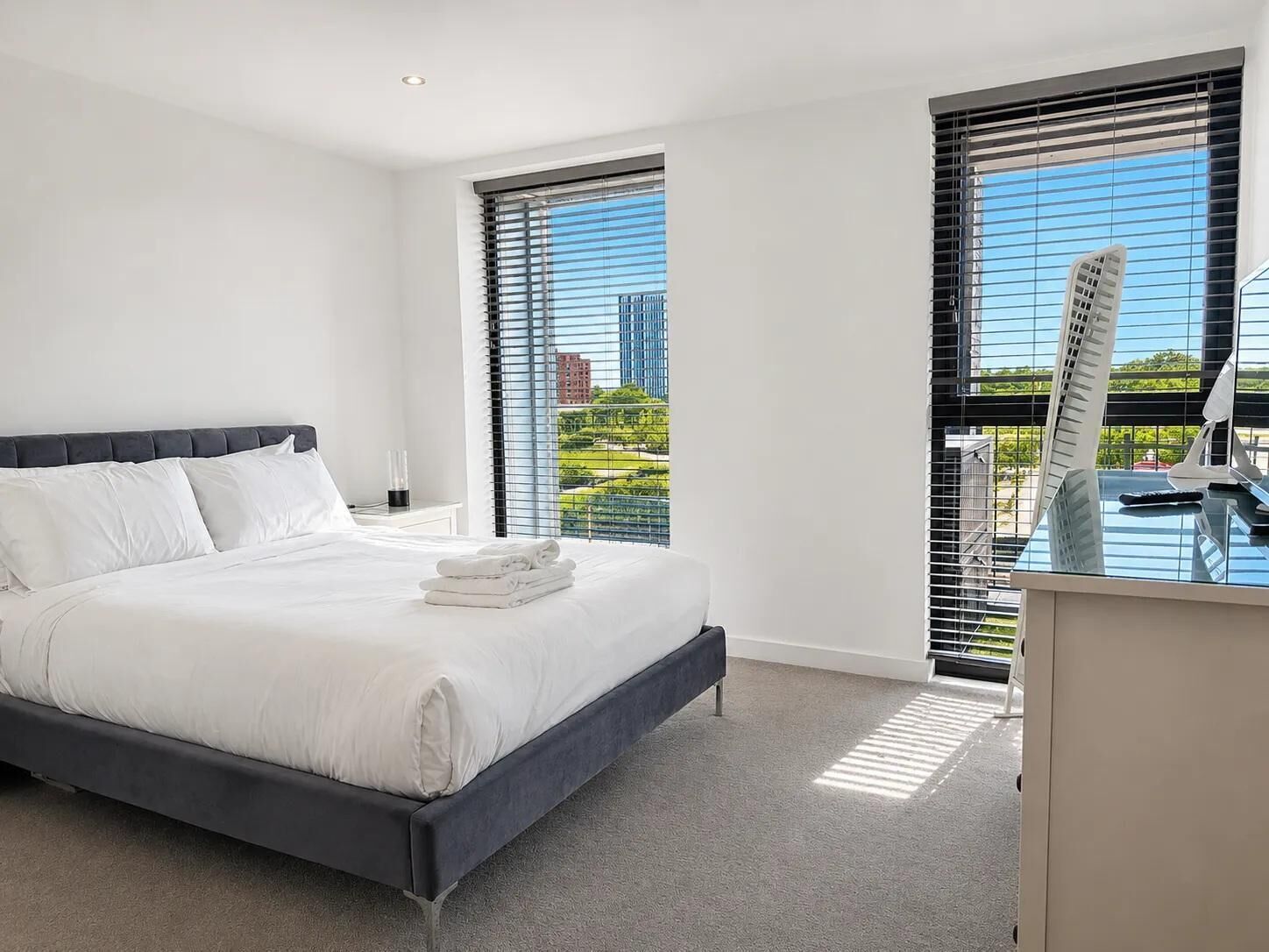 Ein modernes Hotelzimmer mit einem großen Bett, einem Schreibtisch mit Stuhl und Blick auf eine Stadtlandschaft durch raumhohe Fenster.