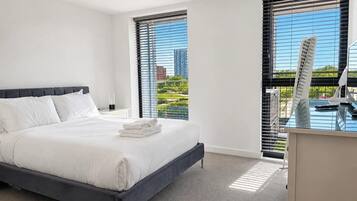 Ein modernes Hotelzimmer mit einem großen Bett, einem Schreibtisch mit Stuhl und Blick auf eine Stadtlandschaft durch raumhohe Fenster.