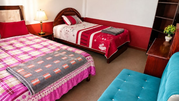 A bedroom with two beds, a bedside table with a lamp, a small wooden nightstand, a blue upholstered chair, and a wooden shelf with a plant.