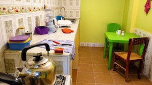 A compact kitchen with a stove, kettle, and various kitchen appliances. There is a dining area with a green table and a chair.