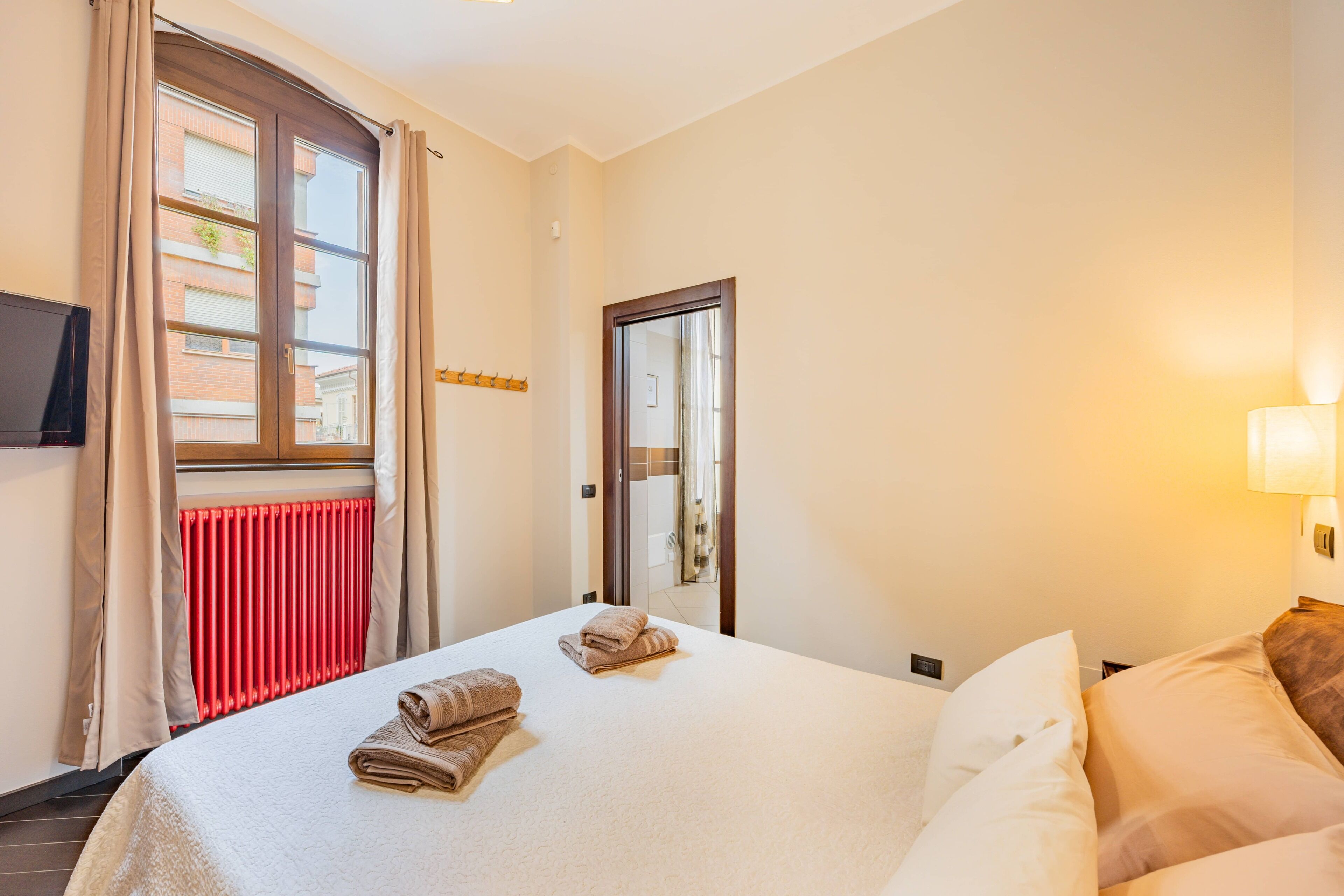 Una camera d'albergo con un letto, una televisione, una finestra con tende, un termosifone e una porta.