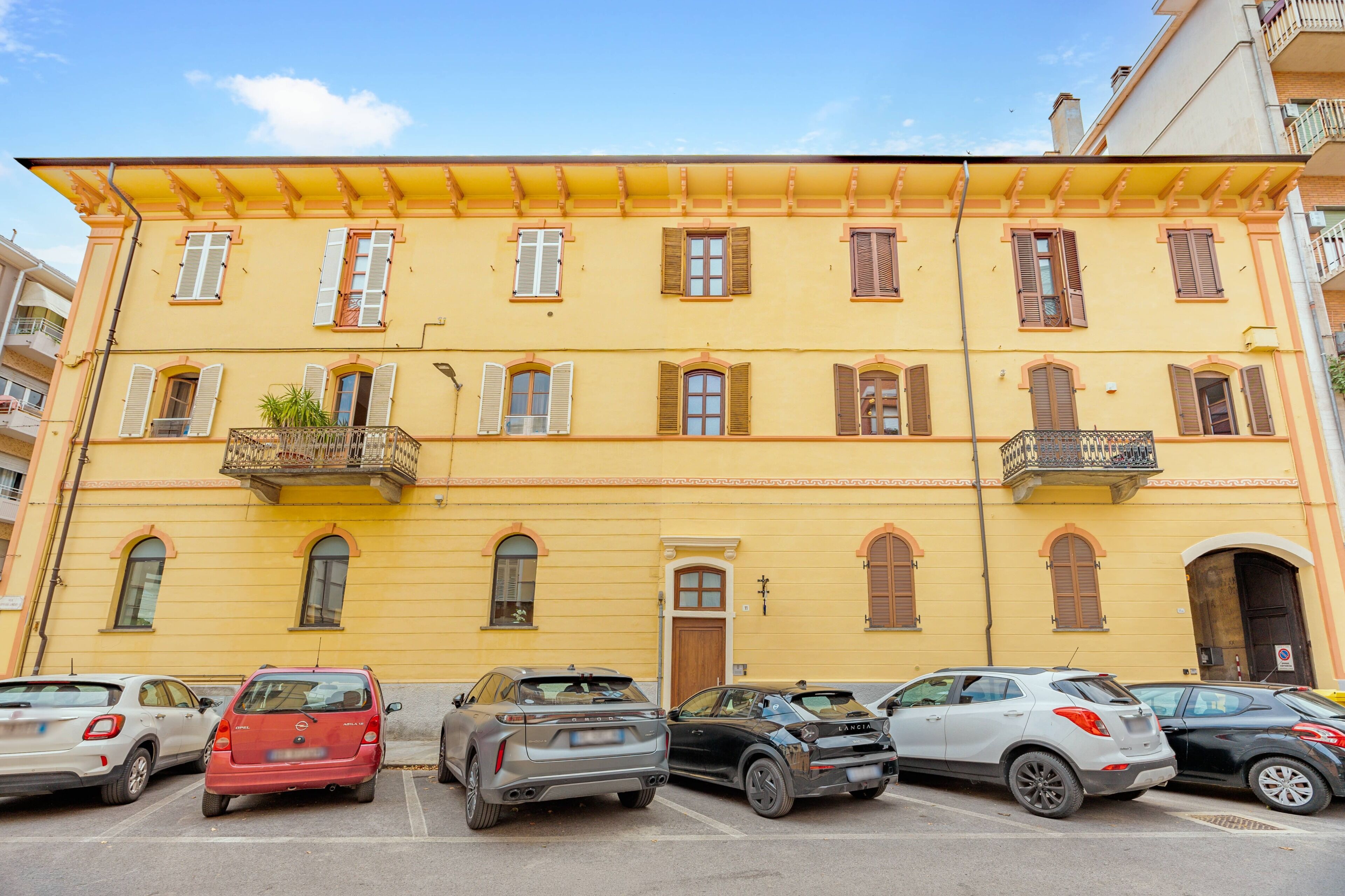 Ein gelbes Gebäude mit zahlreichen Fenstern und Balkonen, Autos vor der Fassade, bewölkter Himmel.