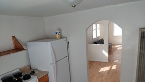 Una cocina compacta con refrigerador, estufa y armarios de madera. Una ventana pequeña proporciona luz natural, y un pasillo conduce a una sala de estar con un sofá.