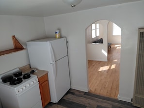 Eine kleine Küche mit Kühlschrank, Herd und Holzschränken. Ein kleines Fenster lässt Tageslicht herein, und ein Flur führt zu einem Wohnbereich mit einer Couch.