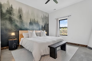 Een slaapkamer met een groot bed, een nachtkastje, een lamp, een bankje, een raam met gordijnen en een bosdecoratie op de muur.