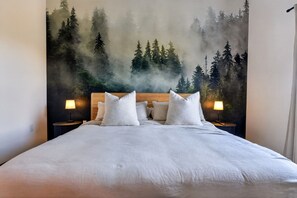 Una cama con ropa blanca, dos lámparas de noche y un mural en la pared con motivo de un bosque.