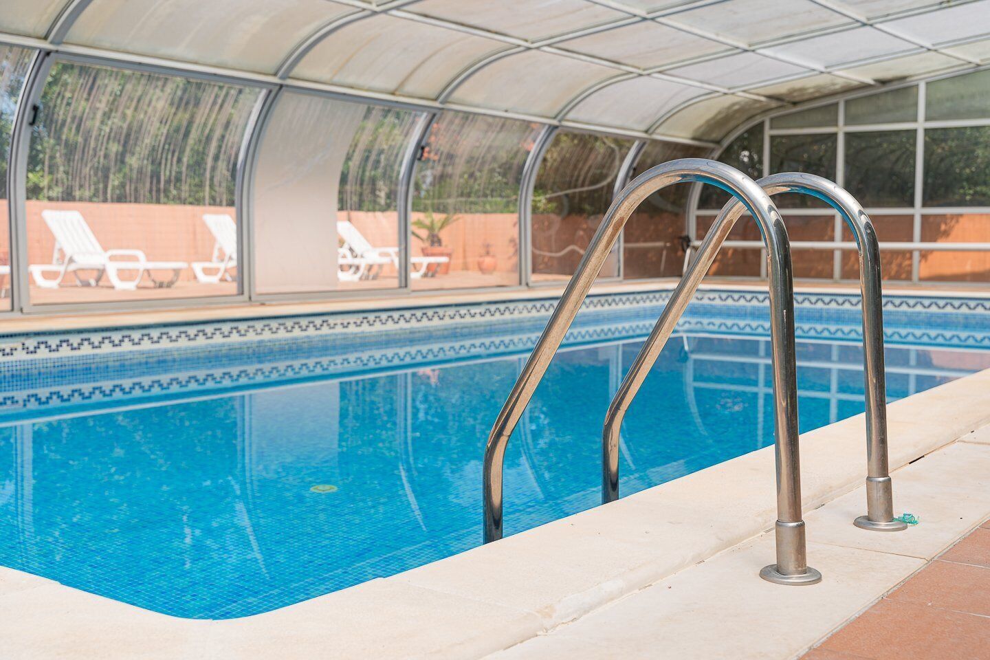 Una piscina interior con paredes de vidrio transparente, escaleras metálicas y borde de cerámica.