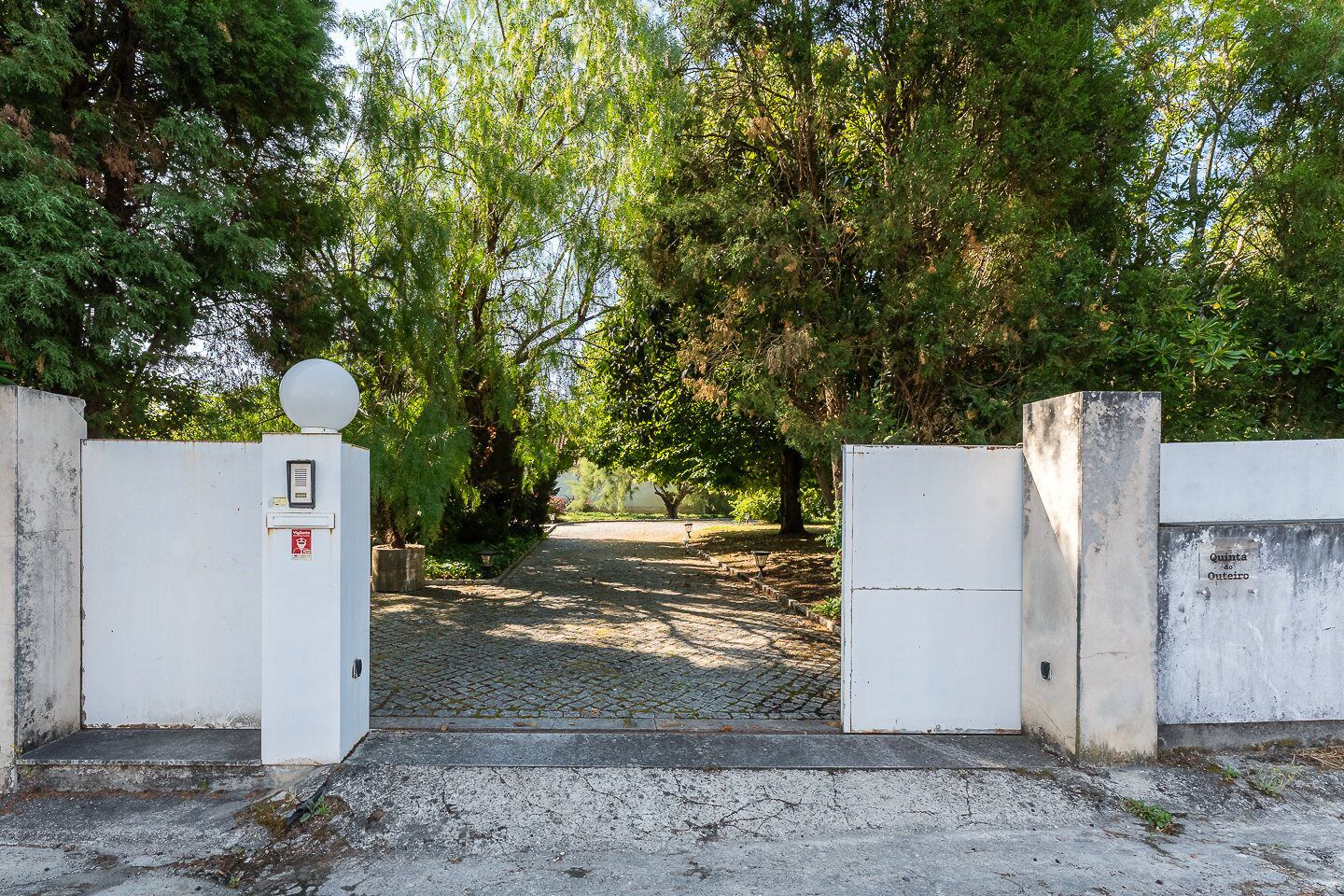 A gated entrance with a white gate, a security camera, and a sign indicating 'Quinta do Outeiro.'