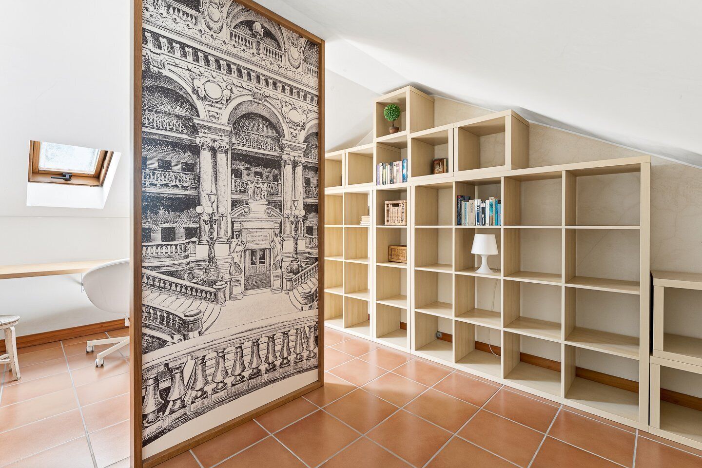 A room with a large framed black and white architectural print, a white desk, a chair, and a wooden bookshelf with books and a lamp.