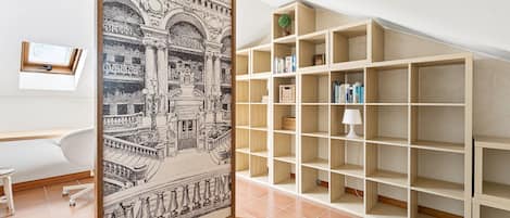 A room with a large framed black and white architectural print, a white desk, a chair, and a wooden bookshelf with books and a lamp.