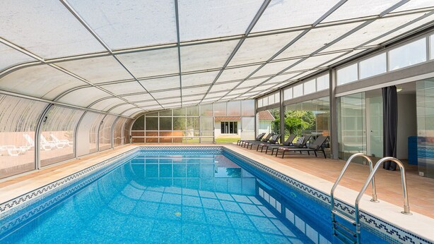 A covered swimming pool with clear glass walls, lounge chairs, and a view of a house. - GuestReady - Quinta do Outeiro (Anadia)