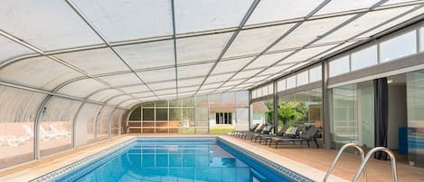 Una piscina techada con paredes de vidrio transparente, sillas de descanso y vistas a una casa.