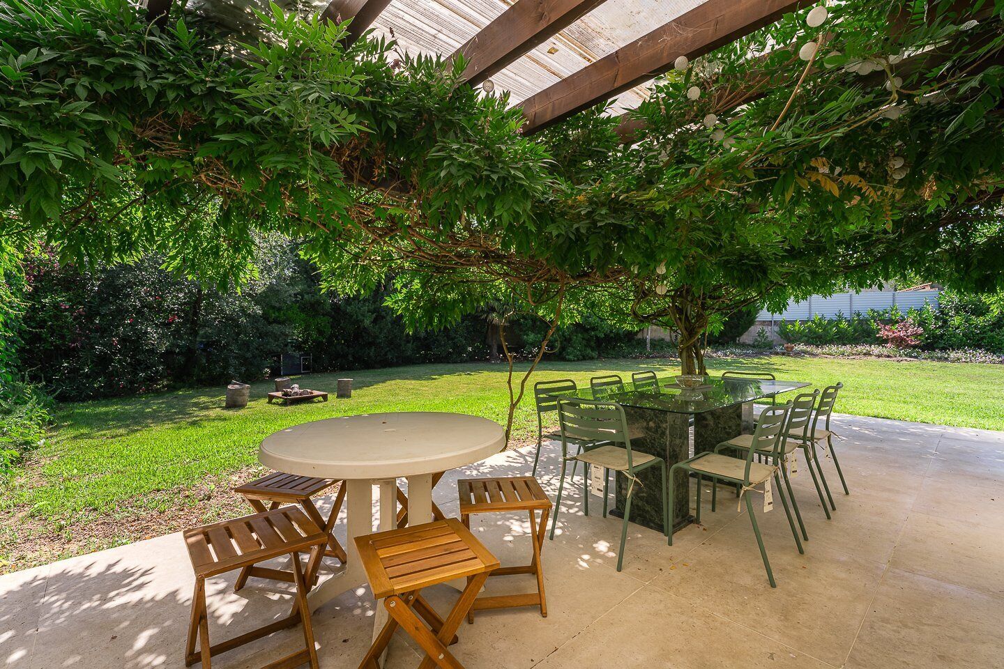 Eine Terrasse im Freien mit einem runden Tisch und Stühlen, umgeben von Grünpflanzen und einer Holzpergola.