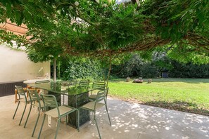 An outdoor patio with a glass table and chairs, surrounded by greenery and a lawn.