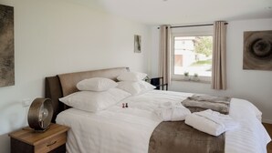 A neatly made bed with white linens, a wooden bedside table with a lamp, and a window with curtains.