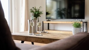 A wooden coffee table with two clear glass bottles and two wine glasses, a potted plant, and a flat-screen TV on a stand.