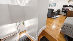 Una escalera conduce a un dormitorio superior con vistas al exterior, un sofá gris, piso de madera y una alfombra pequeña.