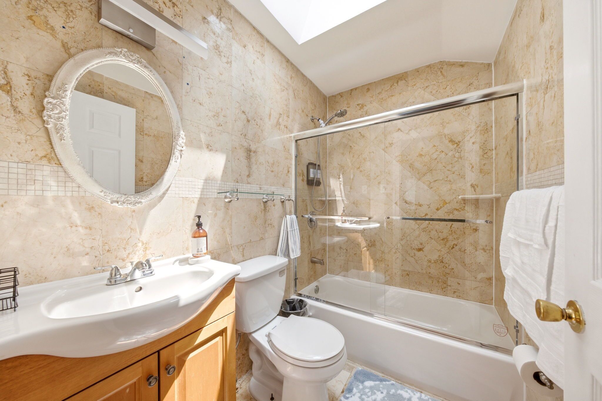 A bathroom with a marble wall, a round mirror, a white sink, a toilet, and a glass-enclosed shower.