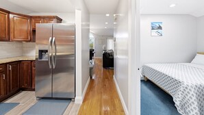 Una cocina moderna con armarios de madera y un refrigerador de acero inoxidable, un pasillo que conduce a un dormitorio con una cama y un cuadro en la pared.