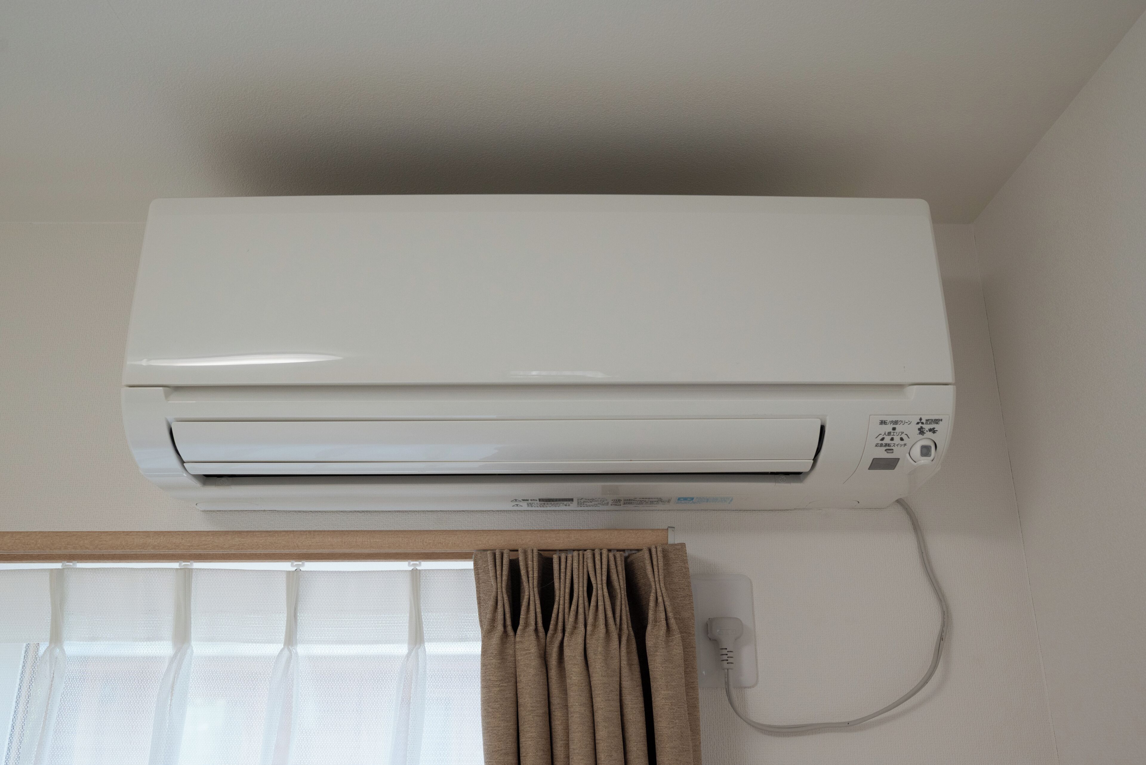 A white air conditioning unit mounted on a wall with a window and curtains below.
